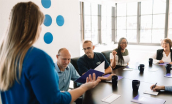 A board room with 5 people, listen to a speaker at the front of the room. The table is filled with Fidelis mugs and folders.