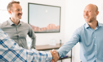 Three business men are in an office, two of them are shaking hands. They are dressed in business casual.