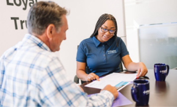 Man wearing a plaid button up and woman in a polo sit at an office table, discussing their paperwork