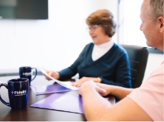 A man wearing a polo and a woman in a sweater sit at an office table and discuss their paperwork. Fidelis mugs and folders are across the table.