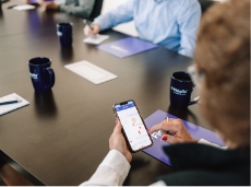 A woman at a conference table looks down at her phone screen while pointing to MedicareCenter.com, which is displayed on the screen.